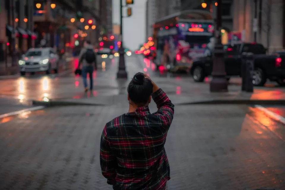 View from behind of person standing on city sidewalk wearing red and black plaid shirt looking across street at dusk.